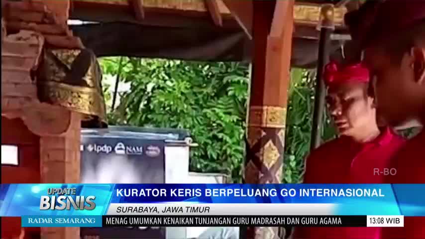 Two men in red are talking, possibly being interviewed, with a Jawa Pos TV graphic at the bottom of the screen. Behind them, a traditional Indonesian structure is visible, with a decorative hanging object.
