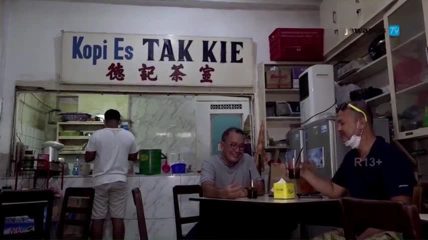 In a small cafe, two men sit at a table, smiling and holding glasses of what looks like iced coffee. Behind the counter, a man in a white shirt is busy preparing something, the scene captured by Jawa Pos TV in Indonesia.
