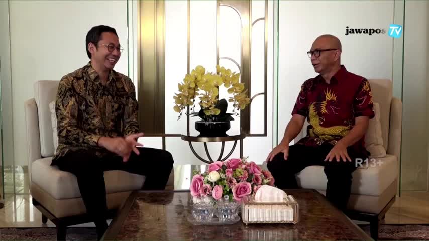 On Jawa Pos TV, two men sit facing each other, engaged in conversation. The man on the left, wearing a patterned batik shirt, gestures as he speaks, and the other, in a red shirt, listens intently.
