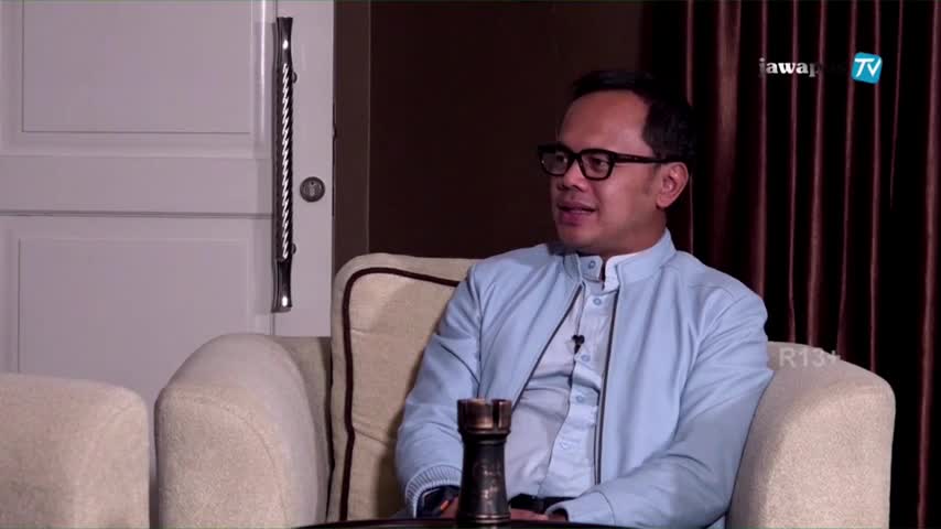 On the Jawa Pos TV set, a man in glasses sits on a cream-colored couch, speaking animatedly. The light blue shirt and the logo in the corner suggest this is an Indonesian broadcast.
