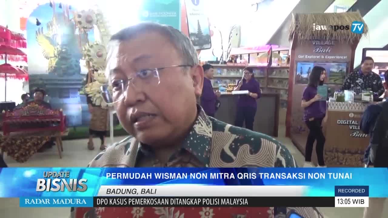 A man in glasses speaks into a microphone, likely for Jawa Pos TV, with a "Digital Village Bali" display behind him. People in purple shirts mill around a booth, and the bottom of the screen displays news headlines from Indonesia.
A man in glasses speaks into a microphone, likely for Jawa Pos TV, with a "Digital Village Bali" display behind him. People in purple shirts mill around a booth, and the bottom of the screen displays news headlines from Indonesia.