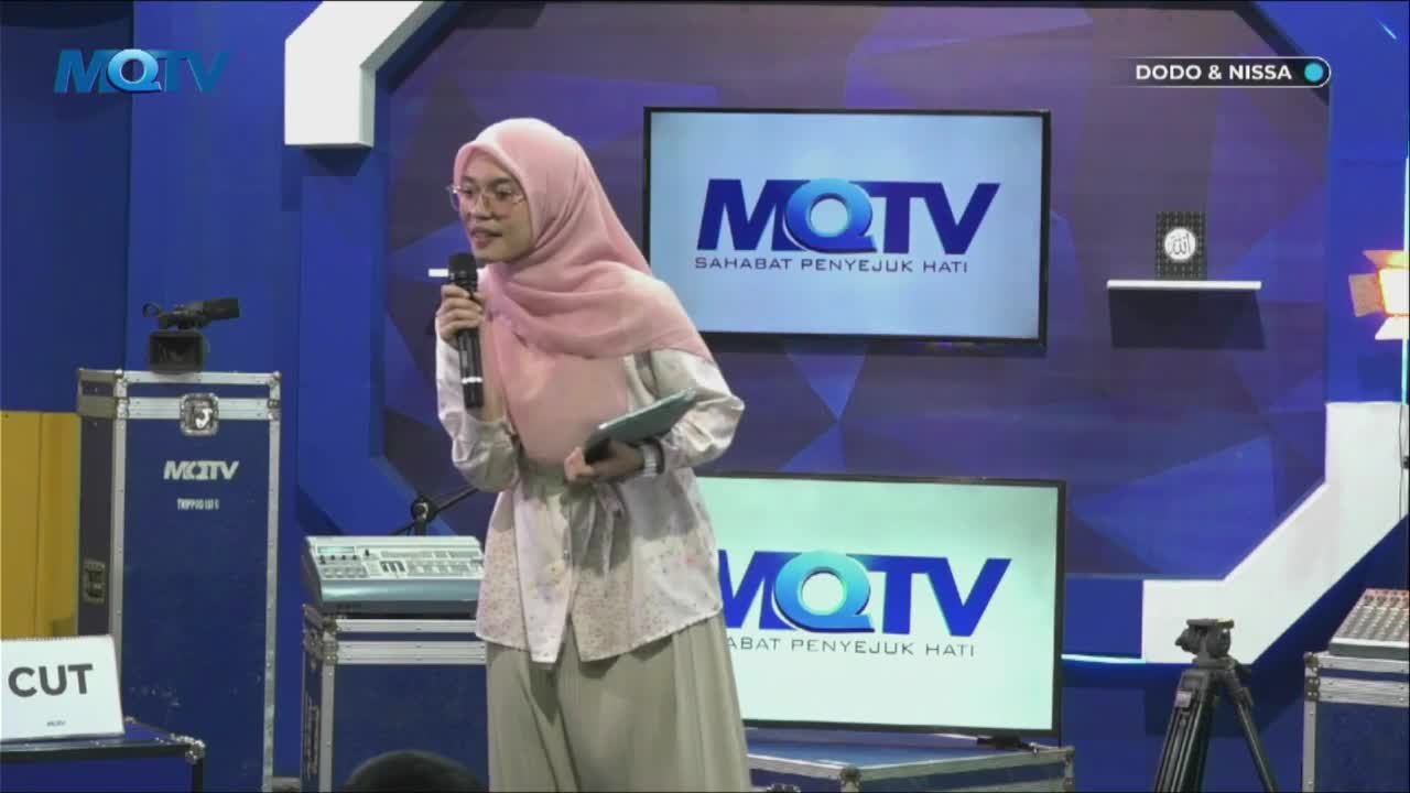 A woman in a pink hijab speaks into a microphone, holding a tablet. Behind her, two screens display the MQTV logo and the show's name.
