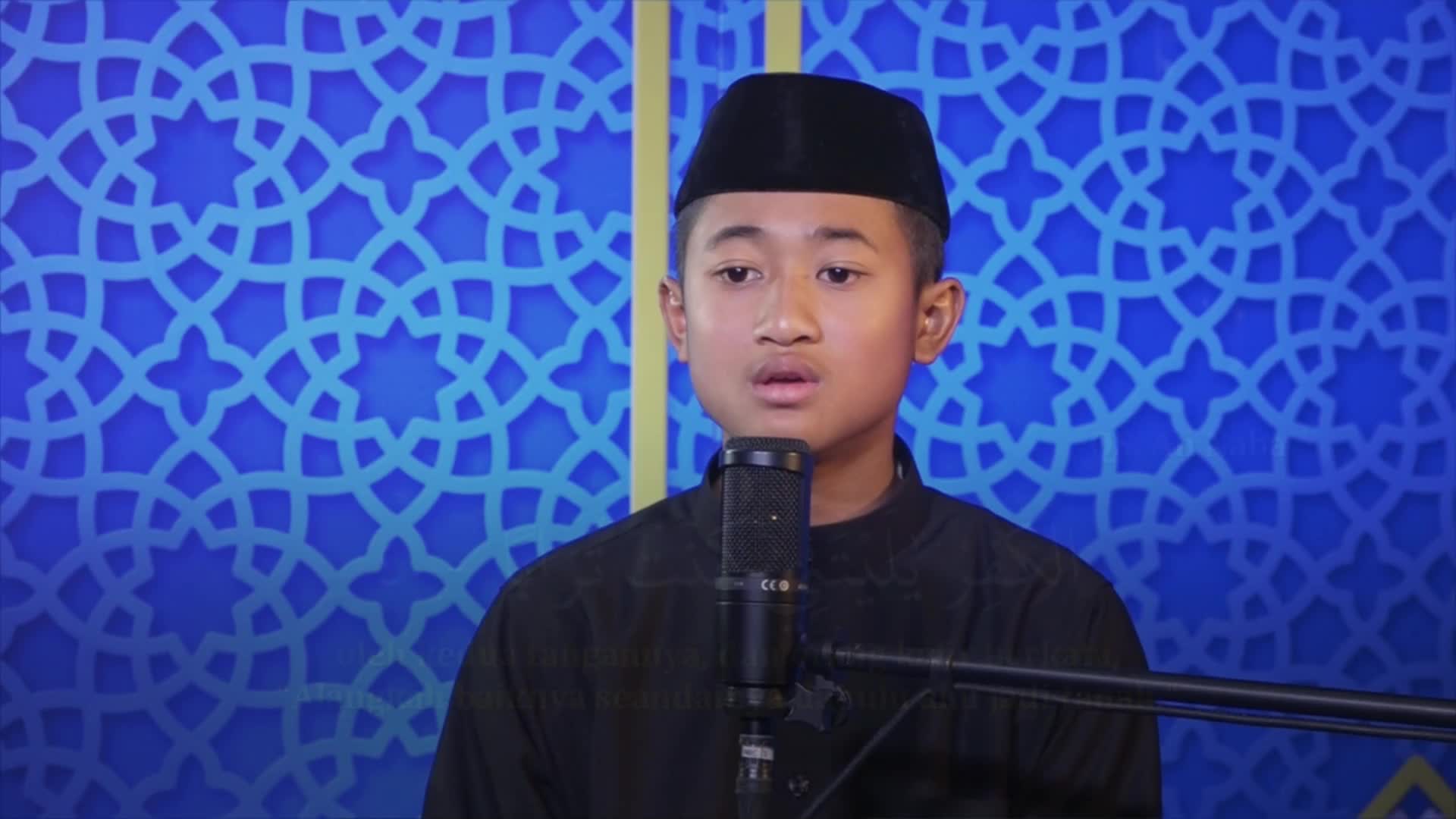 A young man in a black cap and shirt stands before a microphone, his mouth open as if reciting. Behind him, a blue backdrop with a repeating geometric pattern fills the frame.
