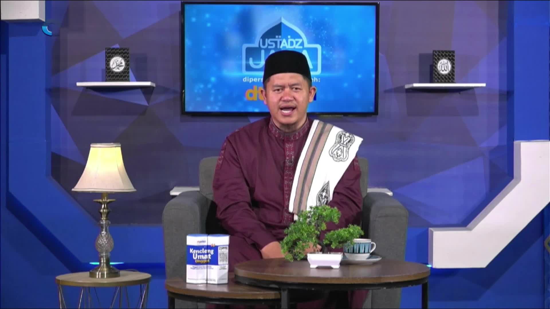 A man in a maroon shirt and black cap sits in a studio, speaking animatedly. Behind him, a screen displays the logo for "Ustadz Jaja" on MQTV.

