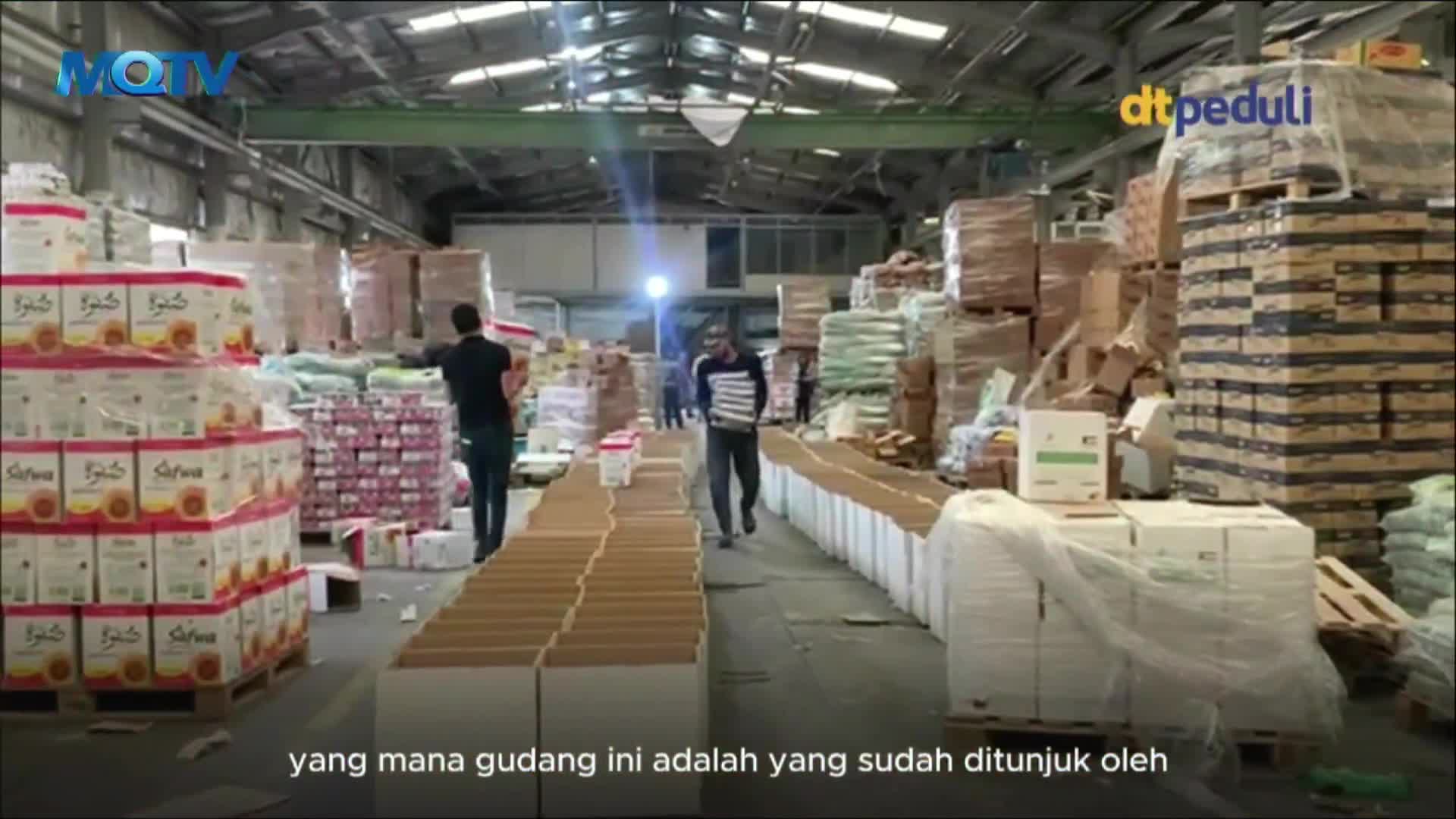 Inside the warehouse, men are carrying stacks of boxes through a maze of cardboard containers and pallets piled high with goods. The MQTV logo is visible, and the scene suggests a distribution center in Indonesia.
