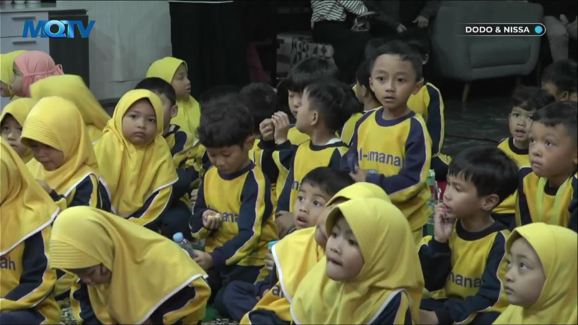 A group of children in matching yellow and navy uniforms sit on the floor, attentively watching something off-camera.  Some of the kids wear yellow headscarves, and the MQTV logo is visible in the corner.
