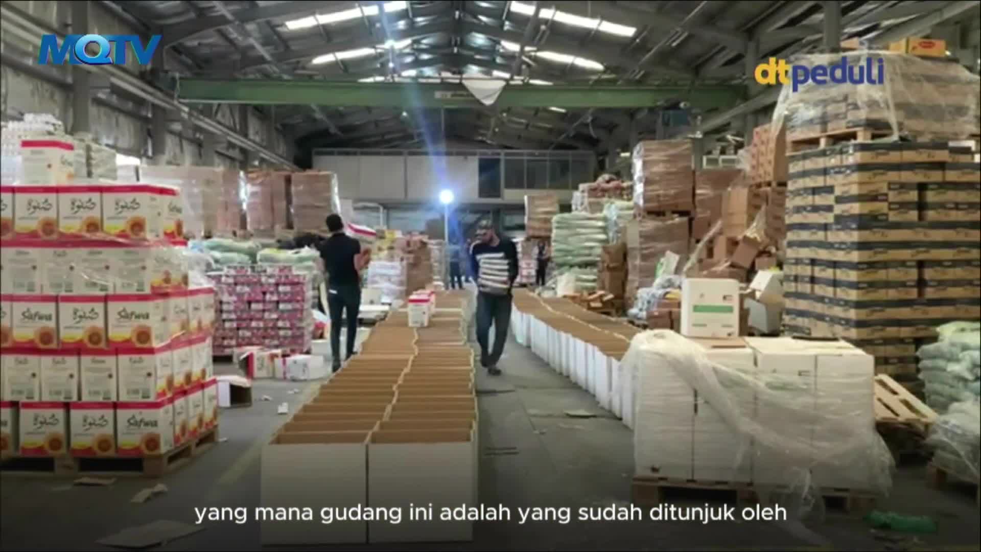 Inside a large warehouse, men are walking between rows of stacked boxes and pallets. The MQTV logo is visible, and the space is filled with supplies.
