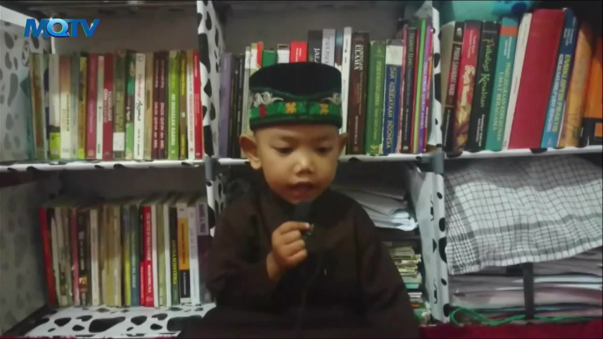 A young boy, wearing a traditional cap and brown robe, speaks into a small microphone. Behind him, a bookshelf is filled with books, and the MQTV logo is visible in the corner.
