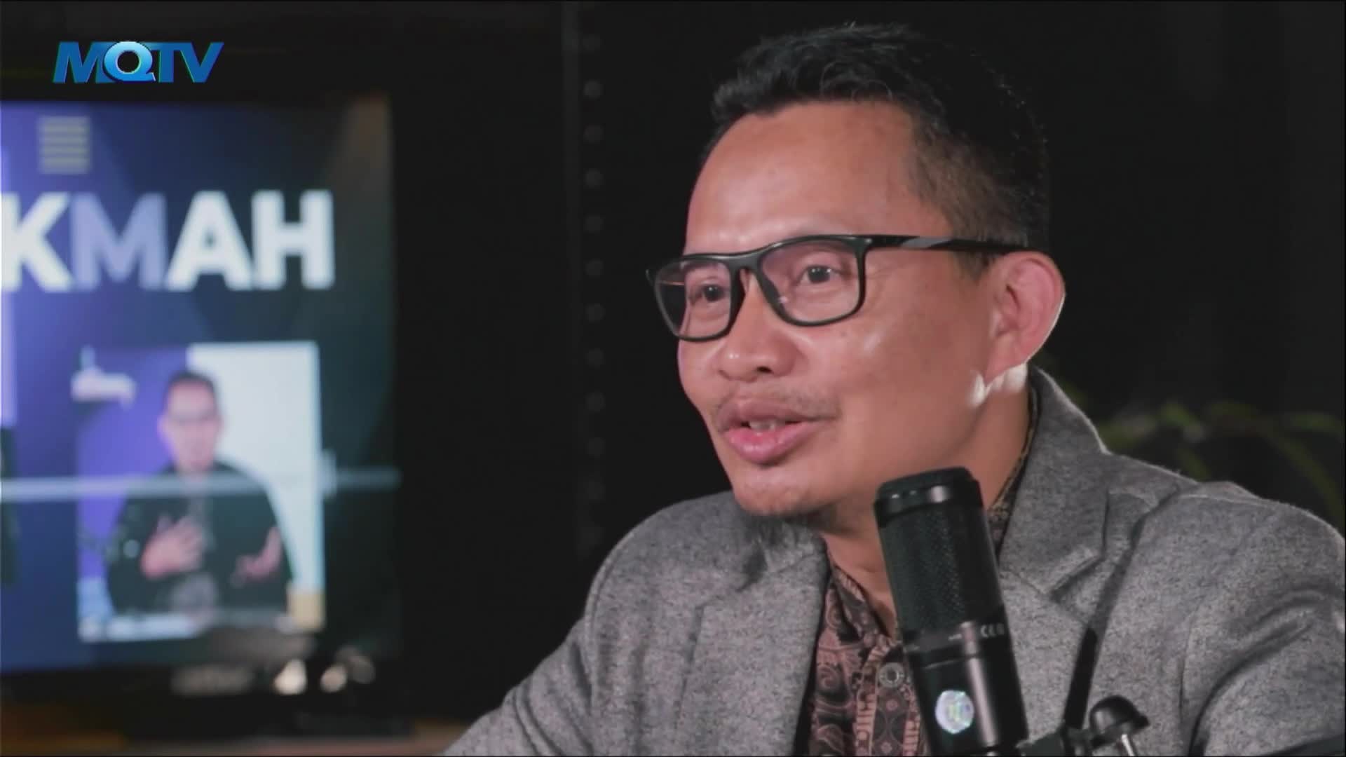 A man in glasses is speaking into a microphone, his expression animated. Behind him, the MQTV logo and the word "KMAH" are visible on a screen, with a blurred image of a person.
