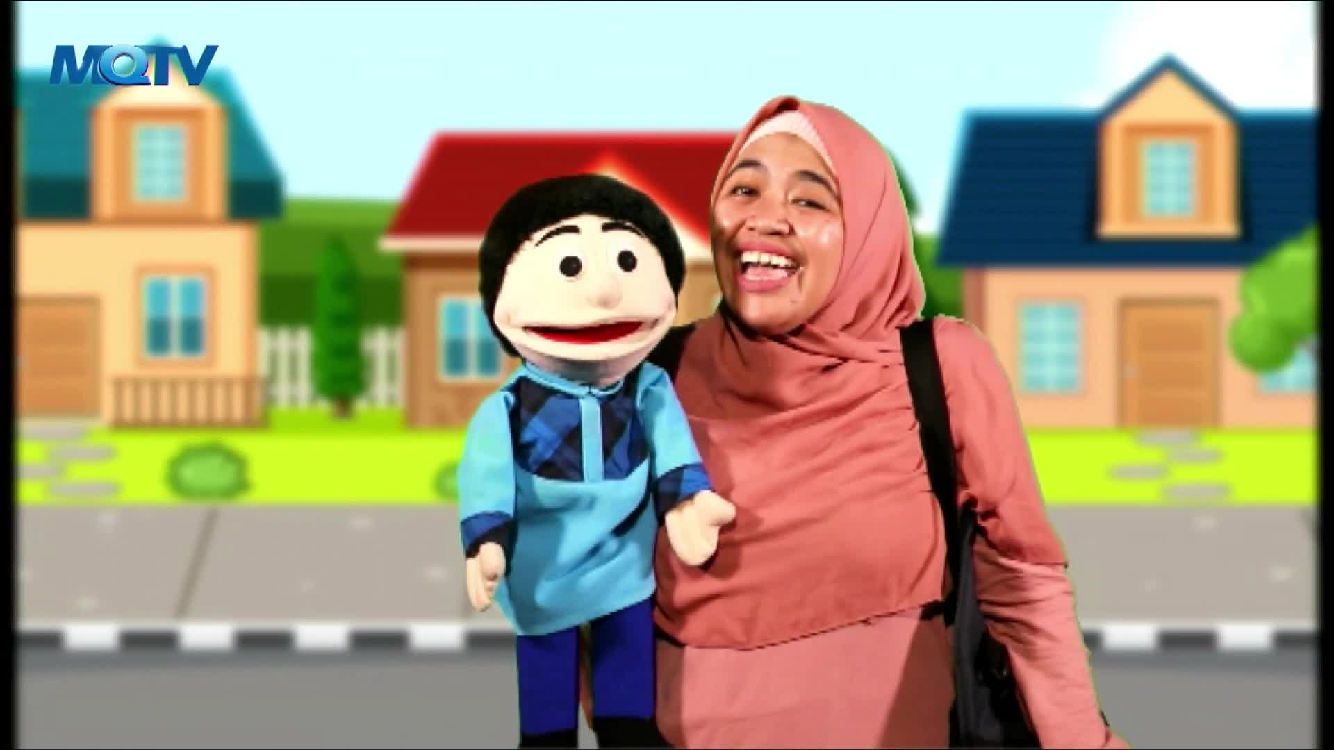 A woman in a pink hijab smiles broadly, holding a puppet dressed in a blue shirt. The background is a cartoon neighborhood scene, likely from an Indonesian MQTV program.
