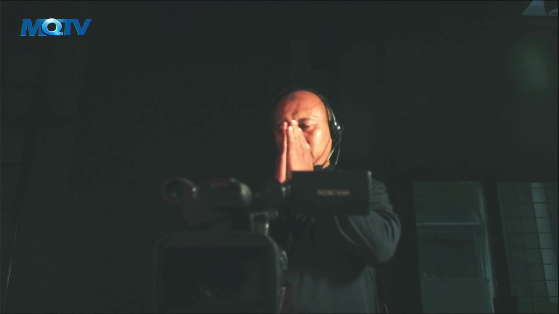 An MQTV cameraman, wearing headphones, stands behind his equipment, his hands clasped in front of his face. He appears to be praying or concentrating intently.
