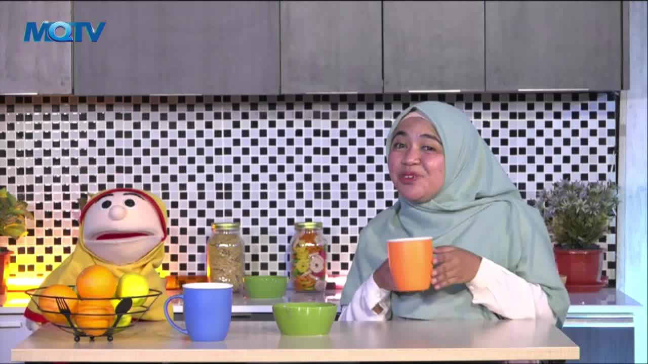 The woman in the hijab holds an orange mug, smiling as she speaks. A puppet sits beside a fruit basket, and the MQTV logo is visible in the background.
