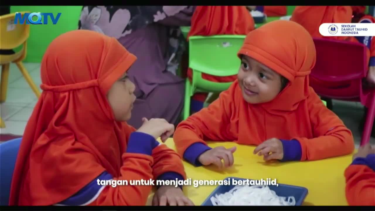 Two small children, dressed in matching orange head coverings and jackets, sit at a yellow table. They are smiling and looking at each other, with a caption in Indonesian at the bottom of the screen.
