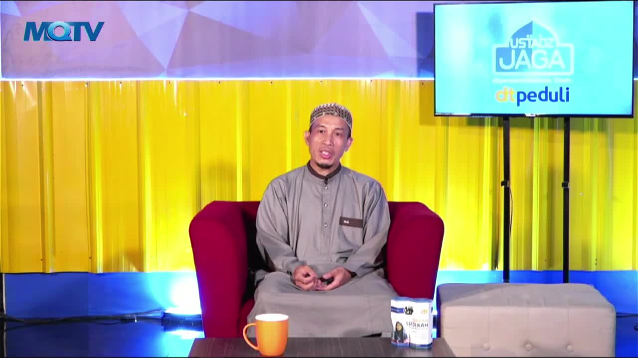 A man in a grey robe sits on a red armchair, speaking directly to the camera. Behind him, a large screen displays a show logo, and the set is illuminated by bright yellow and blue lights.
