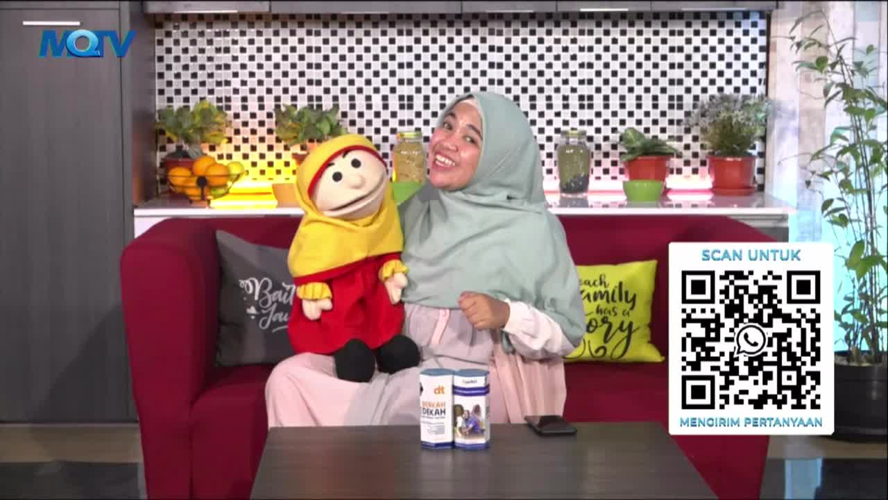 On an MQTV set, a woman in a hijab smiles, gesturing with her hand toward the camera. Beside her, a puppet in a yellow hood and red outfit sits on a red couch.
