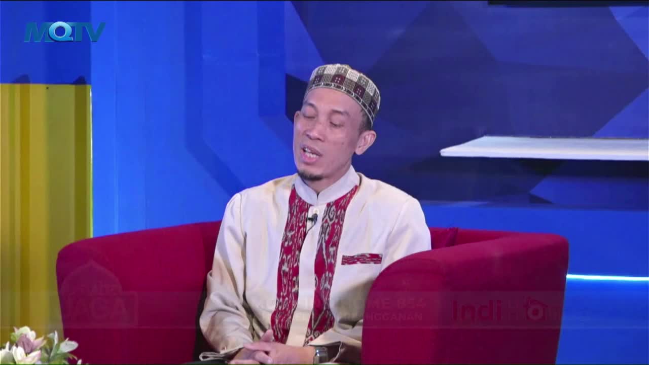 A man in a patterned cap and cream-colored shirt sits in a red armchair, speaking animatedly. The MQTV logo sits in the upper-left corner, and the background is a simple blue and yellow.
