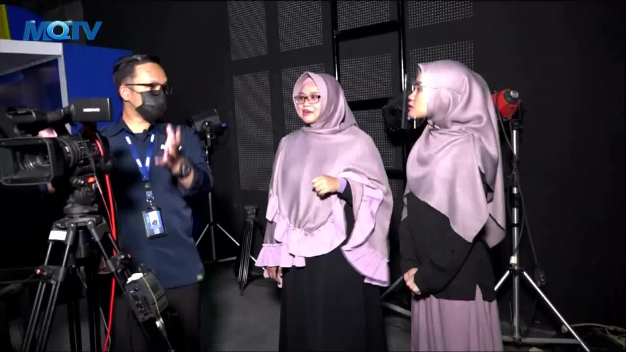 A man in a mask gestures with his hands, explaining something to two women wearing hijabs in an MQTV studio. The women stand still, listening, as a camera on a tripod points toward them.

