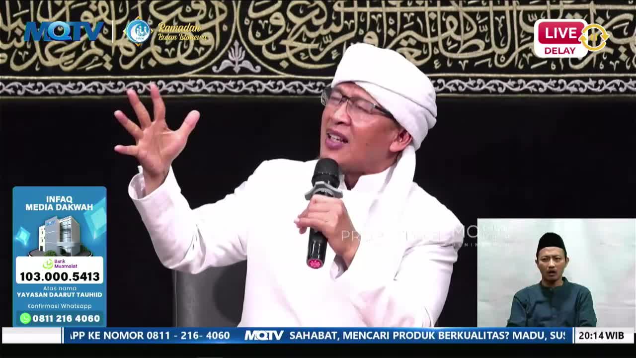 A man in a white robe and turban speaks into a microphone, gesturing with his left hand. On a smaller screen to the right, another man in a dark shirt and peci listens intently.