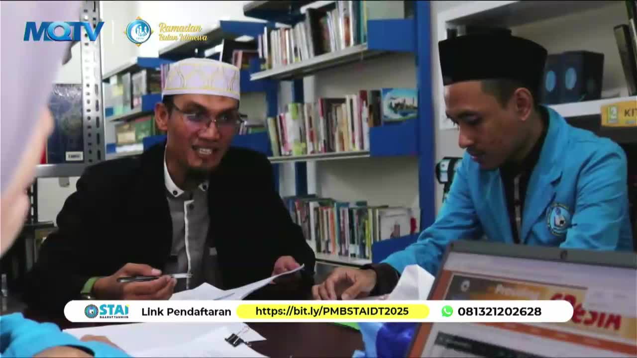 A man in a black jacket and peci gestures with a pen while talking to another man in a blue jacket. Bookshelves filled with volumes line the background of this room.
