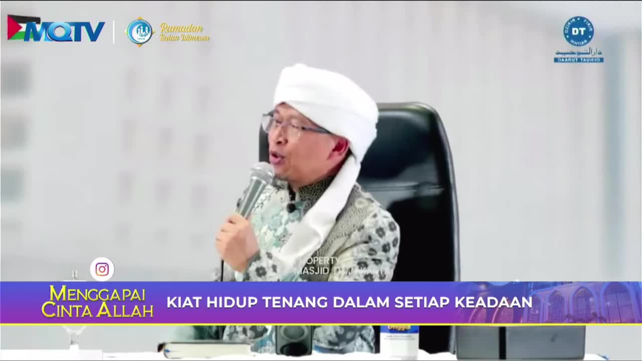 A man in a white turban and patterned shirt speaks into a microphone, his glasses reflecting the light. The MQTV logo is visible in the upper left corner, and Indonesian text scrolls across the bottom of the screen.