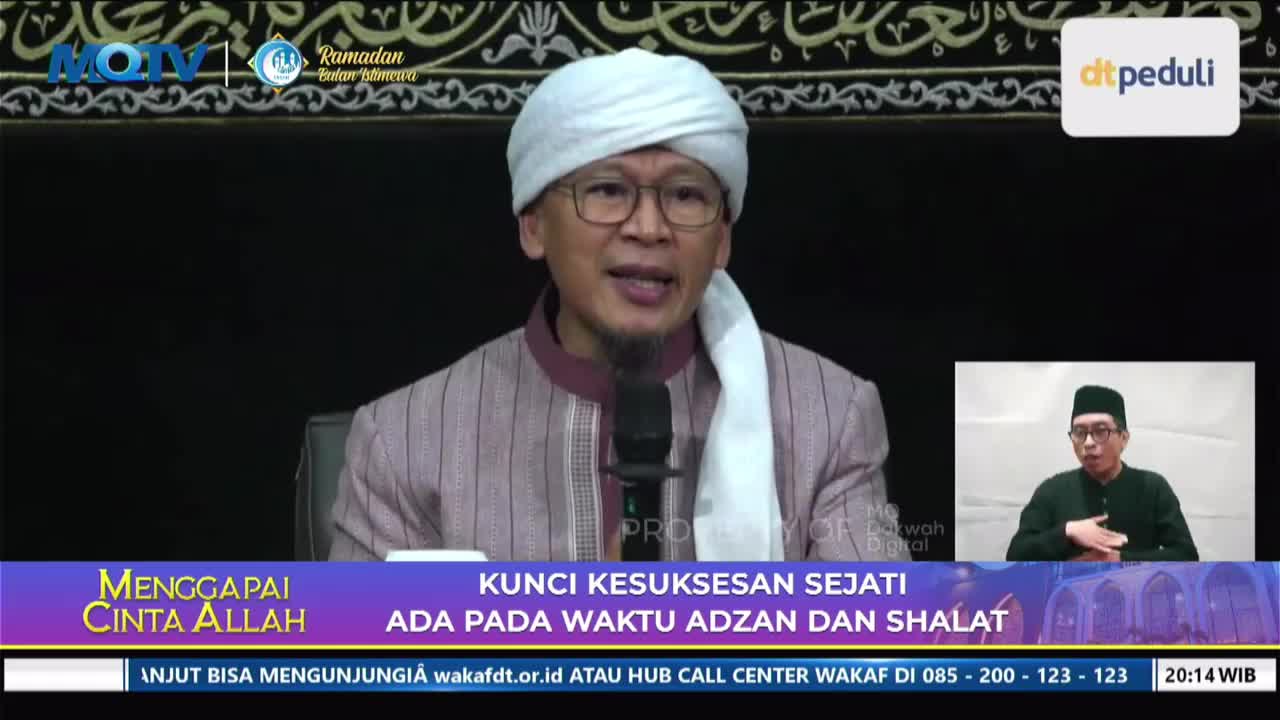 A man in a white turban and striped robe speaks into a microphone, his glasses reflecting the light. A smaller screen in the corner shows another man in a black cap and shirt gesturing with his hand.