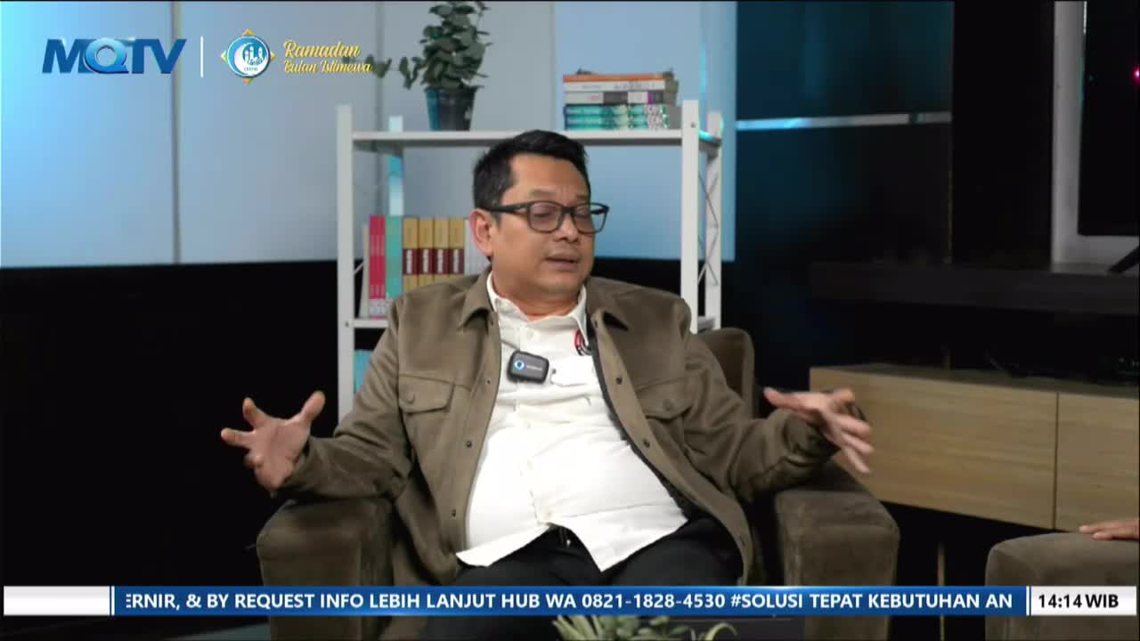 A man gestures with both hands while speaking on a talk show for MQTV. Bookshelves stand behind him, filled with colorful spines.