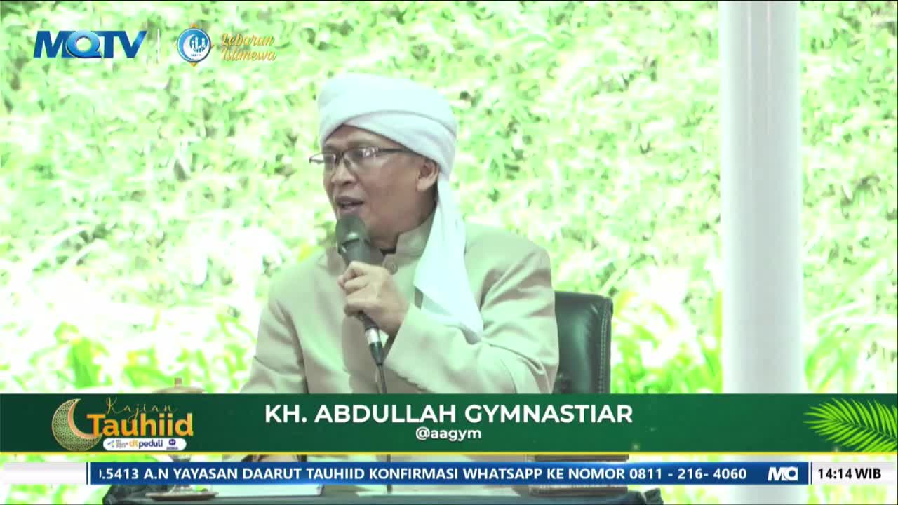 A man in a white turban and glasses speaks into a microphone, his mouth open mid-sentence. He wears a light brown jacket over a white garment, seated in a dark chair.