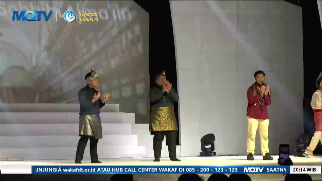 Three men stand on a stage, their hands clasped in front of them. The man in the center wears a gold patterned sarong over black pants. A screen behind them displays the MQTV logo and the words "Lebaran Istimewa."