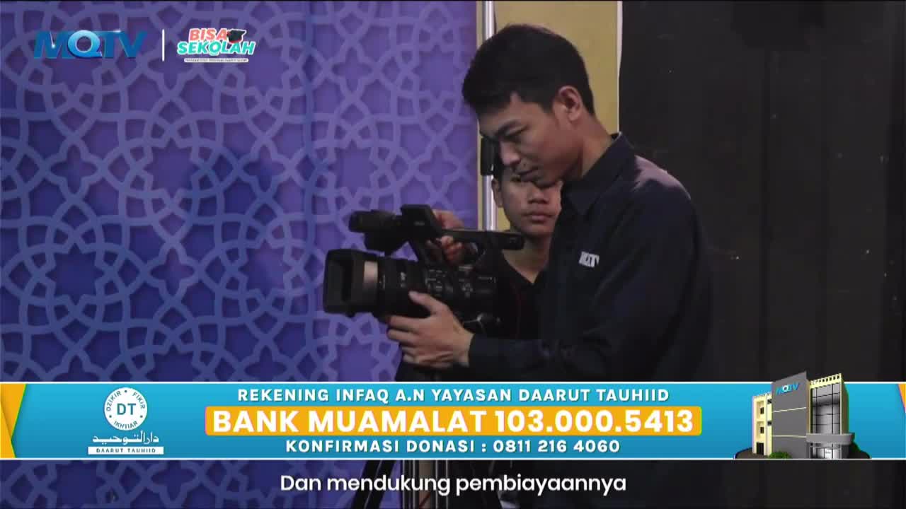 A cameraman adjusts his professional video camera, with another person standing behind him. The MQTV logo and "Bisa Sekolah" text are visible on a patterned backdrop.