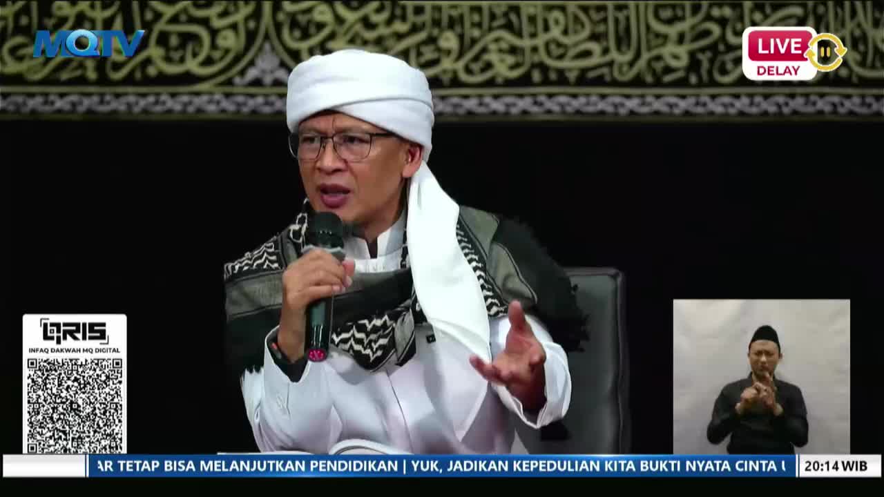 A man in a white turban and traditional clothing speaks into a microphone, gesturing with his hands. In a small box on the right, another man signs what is being said.