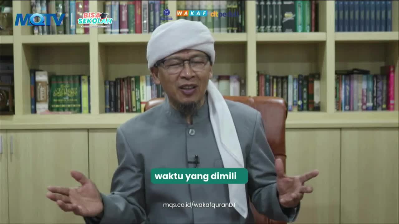 A man in a grey suit and white head covering speaks with animated hands, his voice likely amplified by a microphone clipped to his lapel. Behind him, bookshelves overflow with volumes, a familiar sight on MQTV programs.