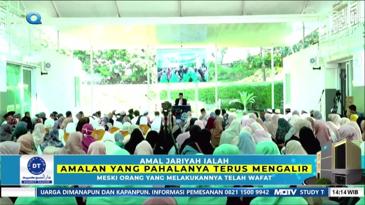 A man in a black cap and robe stands at a podium, addressing a large crowd seated on the floor. The audience, mostly women in colorful hijabs, faces the speaker in a bright, open-air venue.