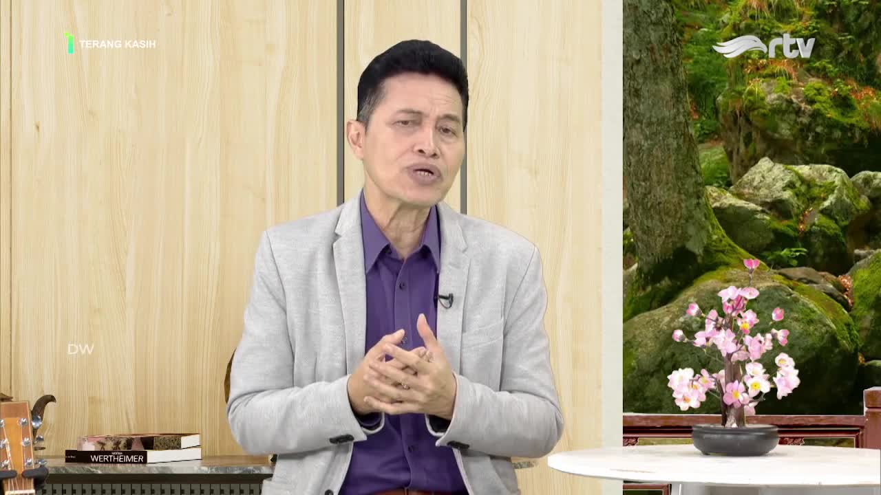 The man in the grey jacket gestures with his hands as he speaks, likely on Rajawali TV in Indonesia. A serene backdrop of mossy rocks and a small flowering plant sits to his right.
