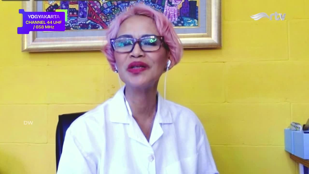 A woman with pink hair and glasses speaks directly into the camera, wearing a white coat, likely for a Rajawali TV broadcast from Yogyakarta. The background is a bright yellow wall, and the screen displays a channel identifier.
