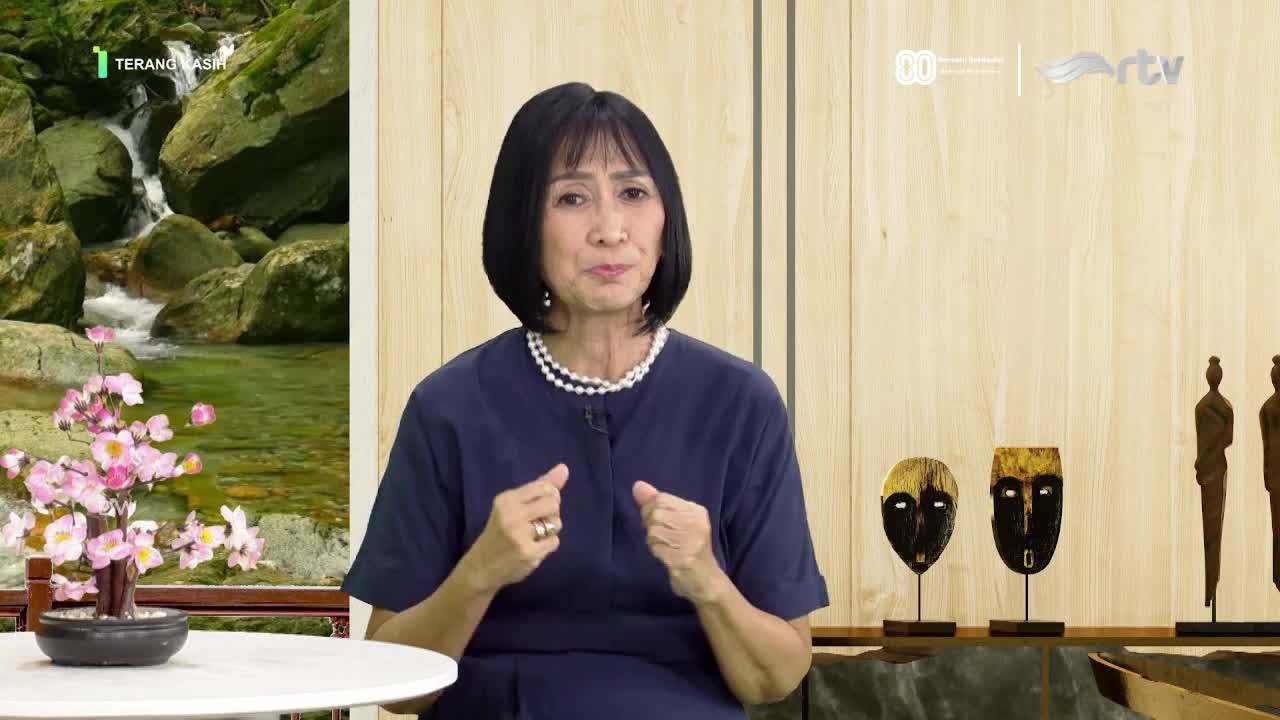 The woman in the blue dress gestures with both hands, speaking directly to the camera. Behind her, a waterfall scene and wooden masks decorate the set, likely for a segment on Rajawali TV in Indonesia.
