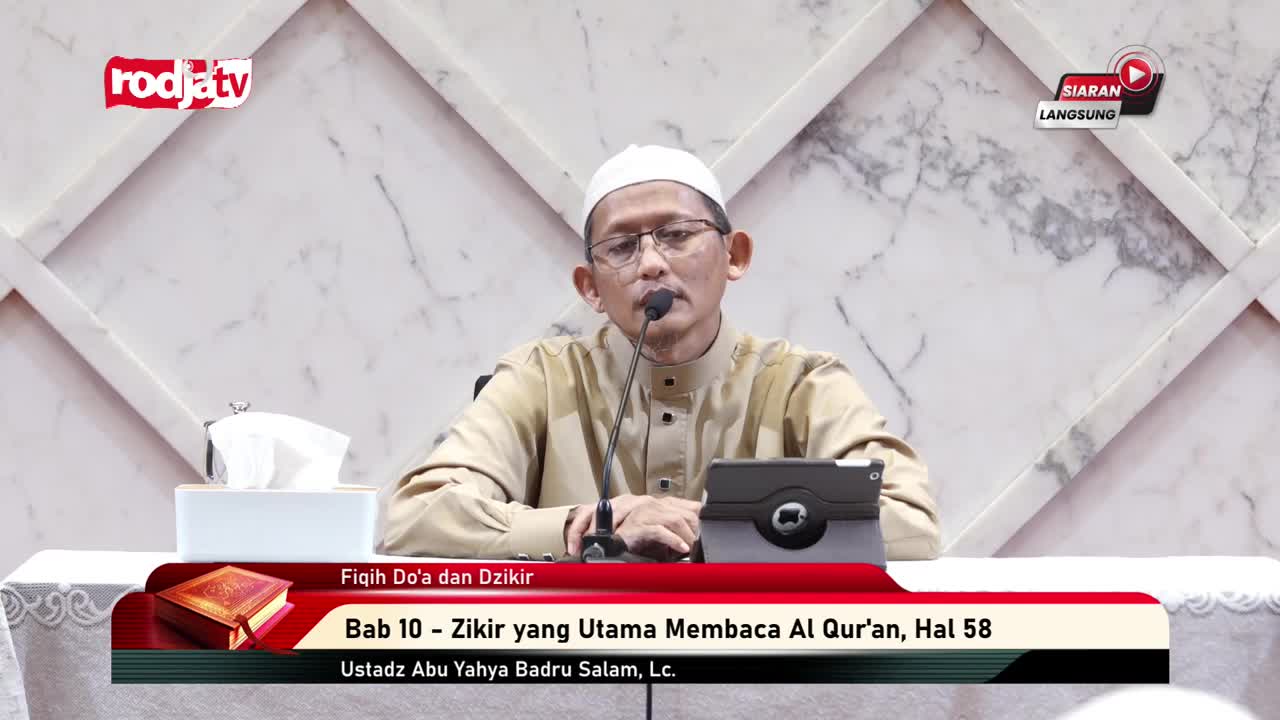 A man in a white cap and glasses speaks into a microphone, likely on Rodja TV in Indonesia. He's seated behind a table with a tablet and a box of tissues, as a banner displays the topic of the broadcast.
