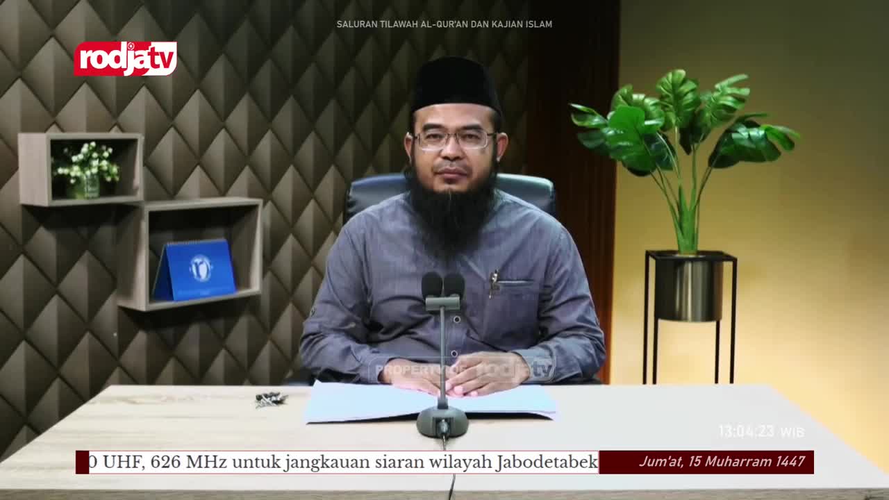 A man with a full beard and glasses sits at a desk, facing the camera on Rodja TV. Two microphones stand in front of him, ready for what looks like a broadcast from Indonesia.

