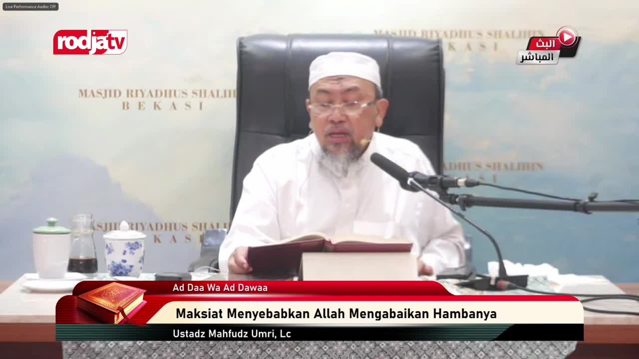 A man in a white shirt and cap sits behind a desk, reading from an open book. He's speaking into a microphone, the Rodja TV logo visible in the corner.
