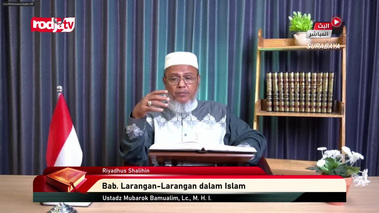 A man in a white cap gestures with his hands as he speaks, a book open before him on a small lectern. The Rodja TV logo is visible, and the Indonesian flag stands to his left.
