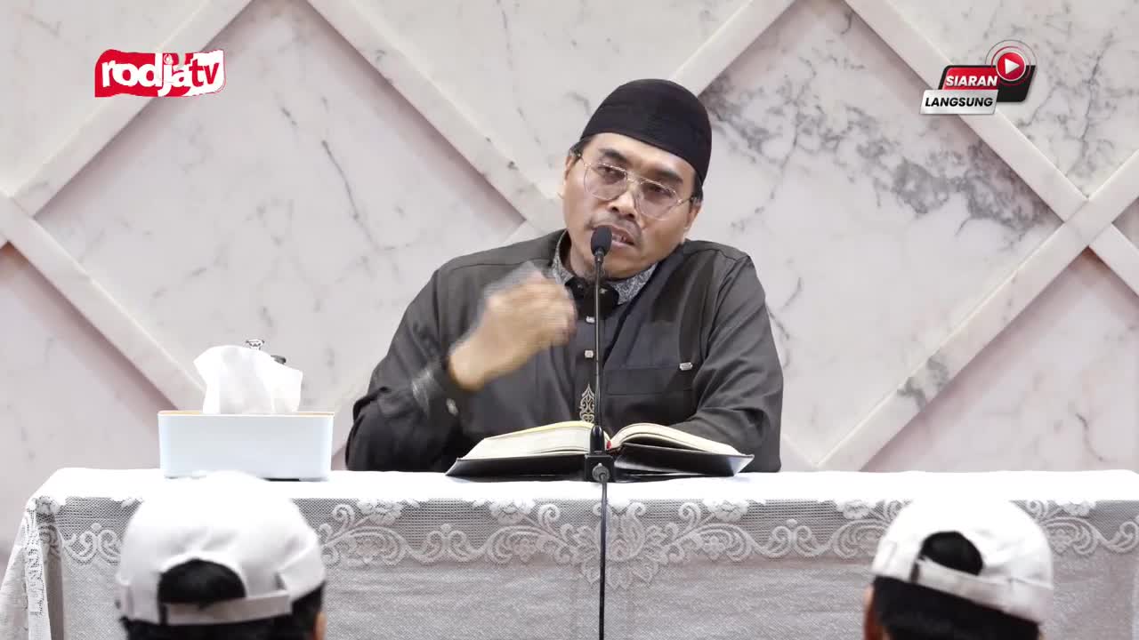 A man in a black cap gestures with his right hand while speaking into a microphone. He's sitting at a table with a book open in front of him, and the Rodja TV logo is visible in the corner.
