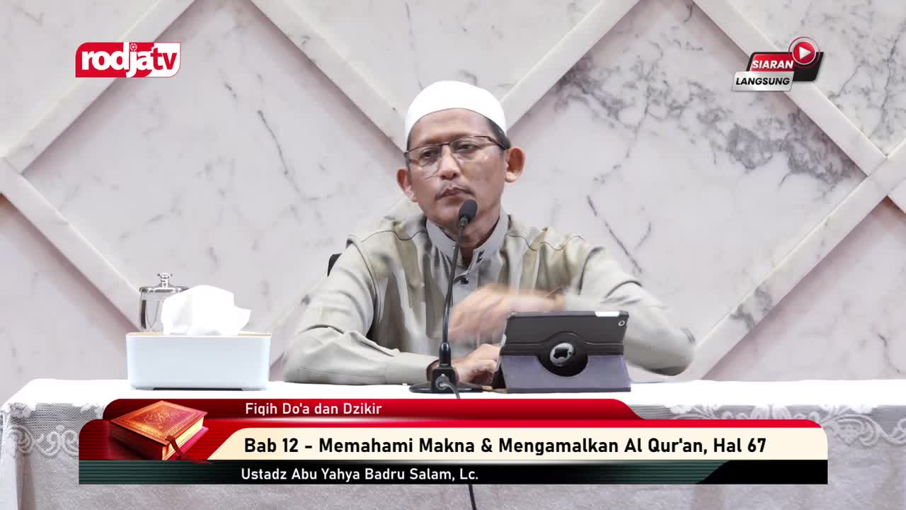 A man with glasses and a white cap speaks into a microphone, likely on Rodja TV in Indonesia. A tablet sits on the table, and the lower third of the screen displays the topic of the discussion.
