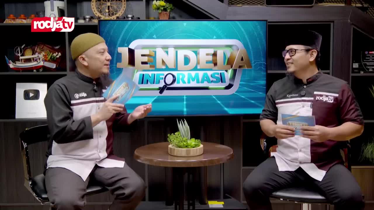 Two men in matching shirts sit facing each other on the set of Rodja TV's "Jendela Informasi" program, each holding a small booklet. The man on the left gestures with his booklet as he speaks, while the other listens attentively.
