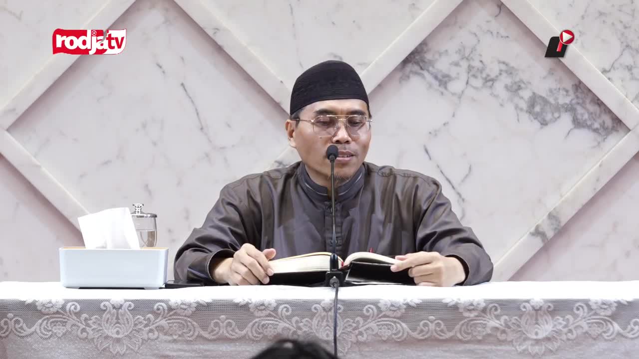A man in a black cap speaks into a microphone, reading from a book open on the table in front of him. The Rodja TV logo is visible in the corner of the screen.
