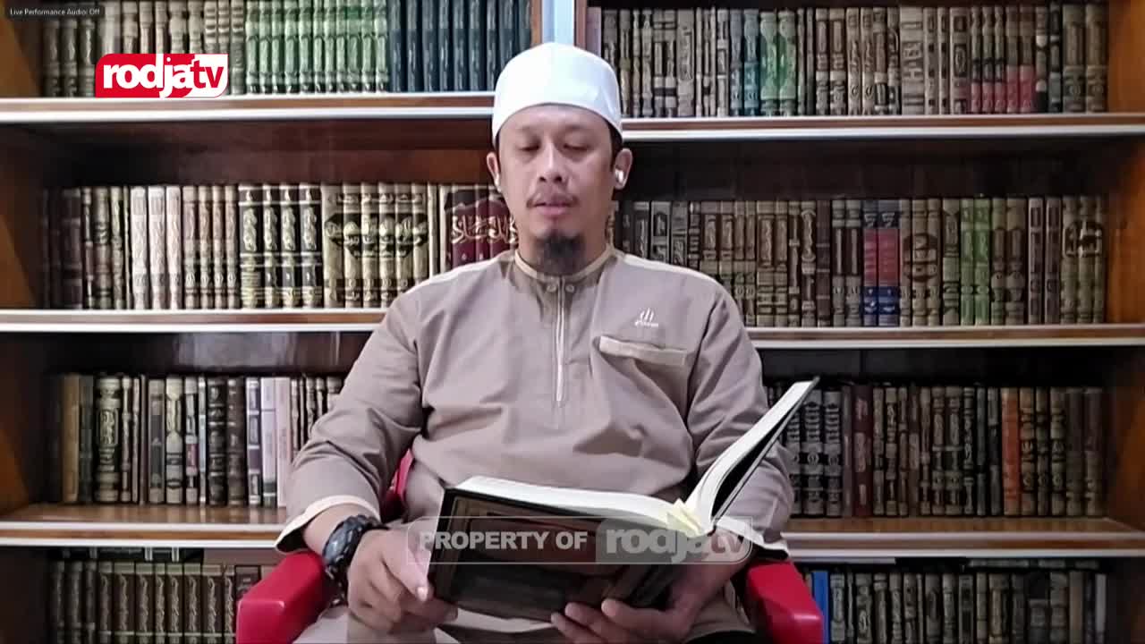 A man in a white cap and tan tunic sits in front of a Rodja TV backdrop, reading from an open book. The camera angle is close, showing his focused expression as he holds the pages.
