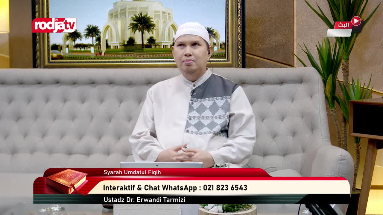 A man in a white cap and shirt sits on a plush couch, speaking on Rodja TV in Indonesia. The broadcast is about Syarah Umdatul Fiqih, with a WhatsApp number displayed for interactive chat.
