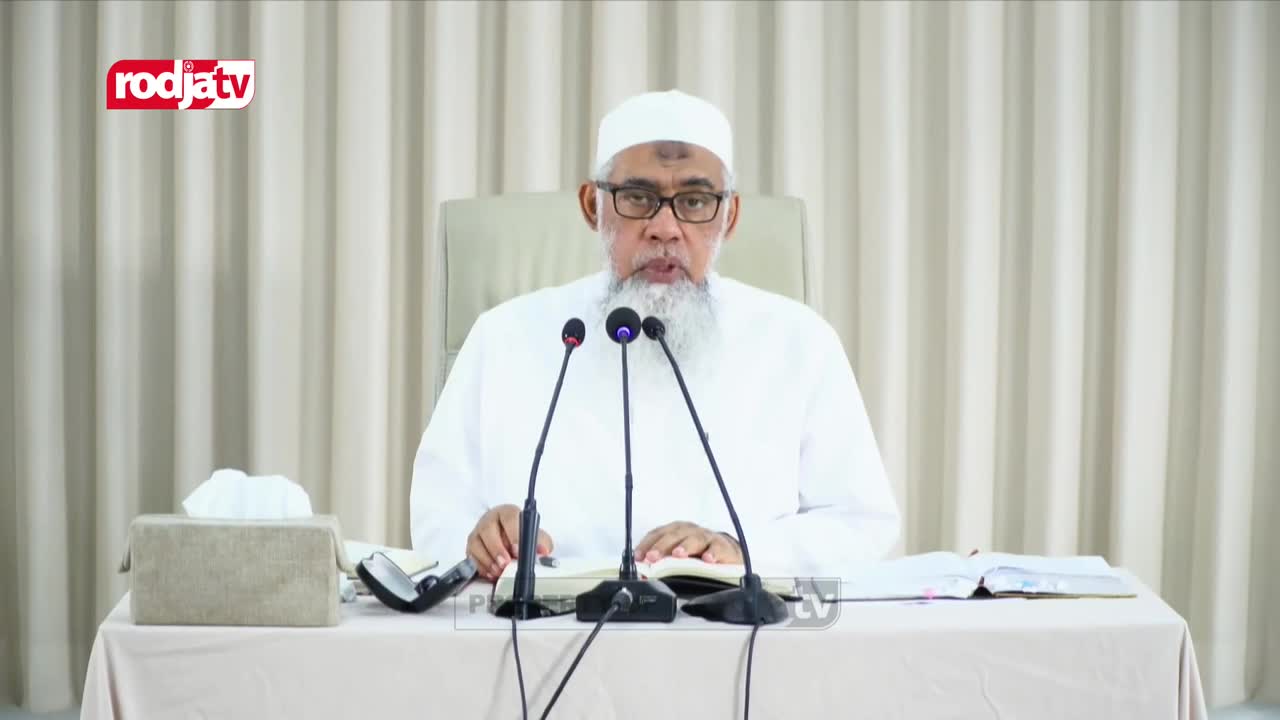 A man in white robes sits at a table, facing the camera with three microphones in front of him. The Rodja TV logo is visible in the top left corner of the screen.
