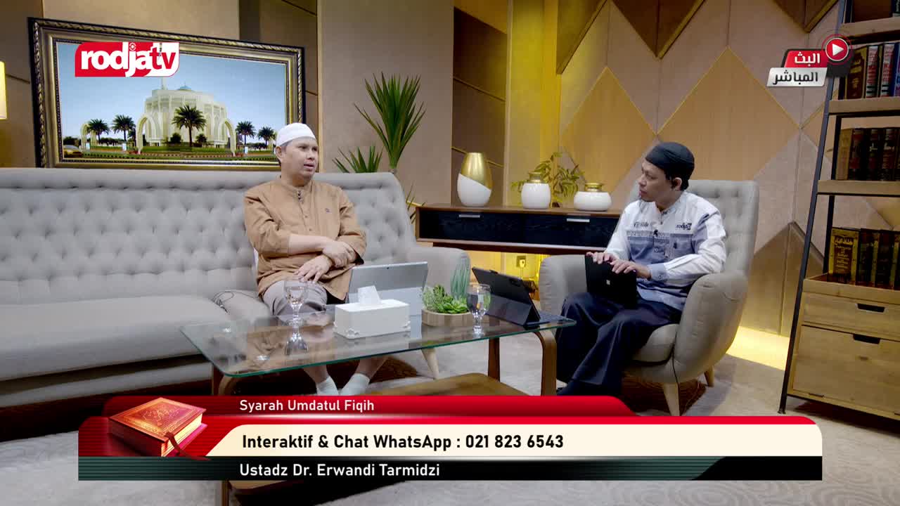 Two men, both wearing white caps, sit facing each other in a studio, likely for Rodja TV in Indonesia. One man, seated on a sofa, gestures with his hands as the other, sitting in an armchair, looks at a laptop.
