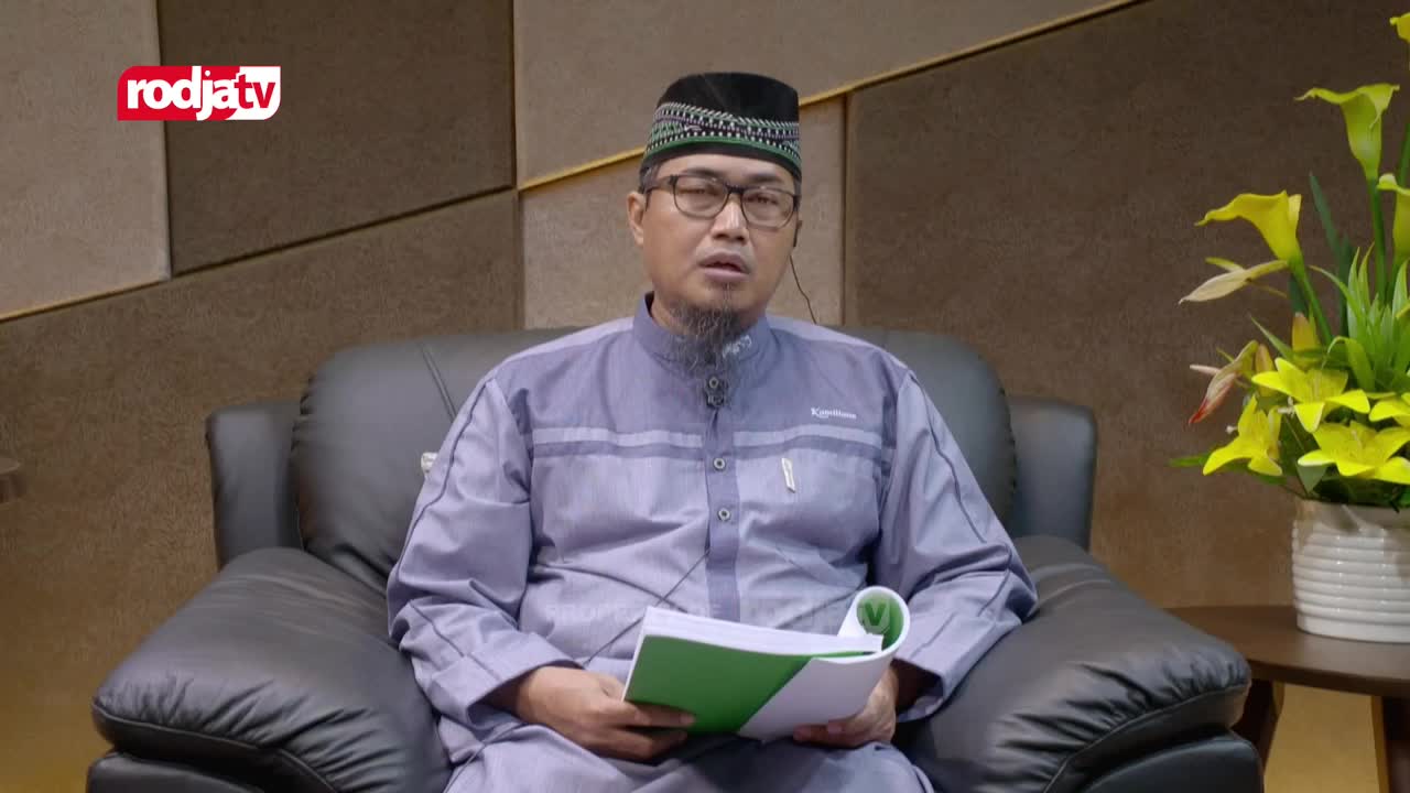 A man wearing glasses and a traditional cap sits in a leather armchair, speaking into a microphone. He's holding a green-covered book, likely for reference, as he addresses the camera on Rodja TV.

