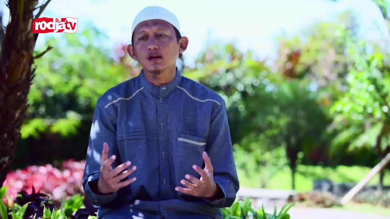 A man in a white cap and blue shirt gestures with his hands as he speaks. The Rodja TV logo is visible in the upper left corner, and the background is a lush green park.
