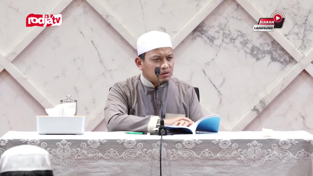 A man in a white cap speaks into a microphone, referencing notes in a blue-covered book. The Rodja TV logo and "Siaran Langsung" (Live Broadcast) are visible, indicating a live program is underway.
